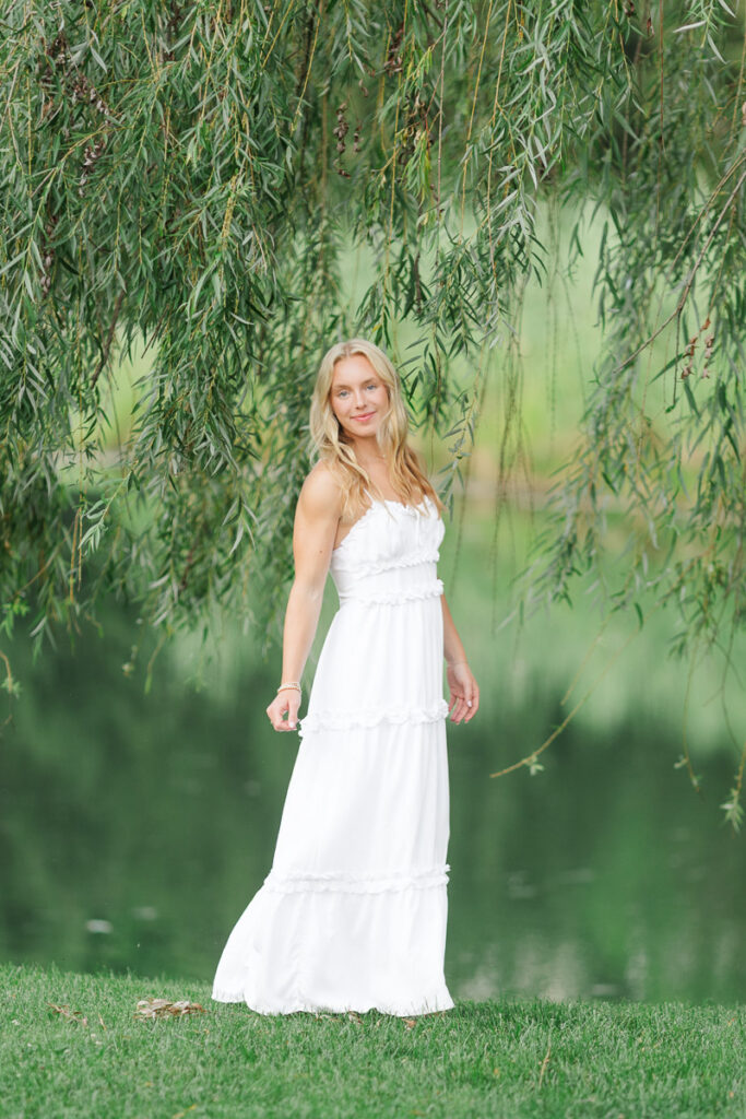 a senior girl standing by a willow tree wearing a white dress