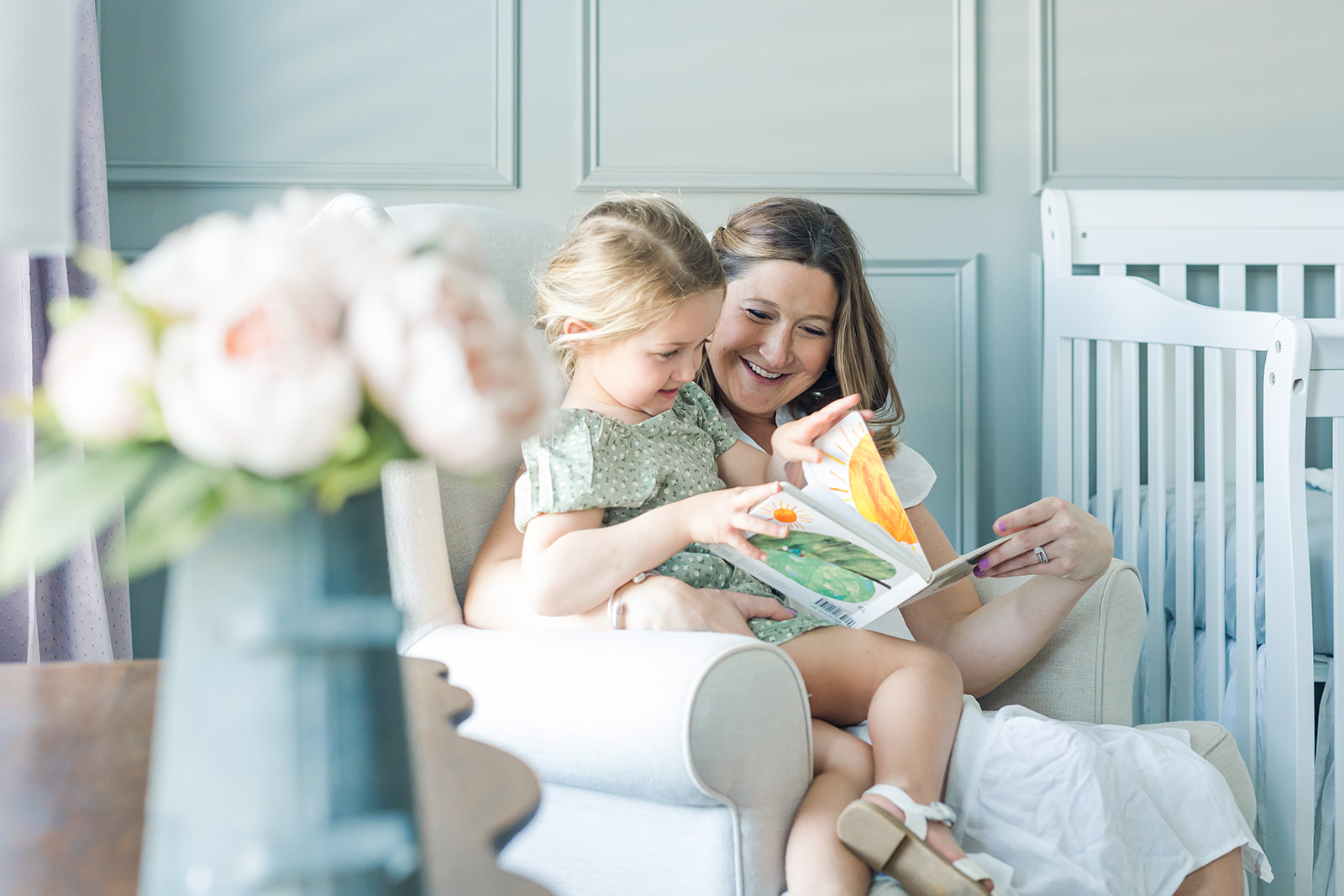 A mother sits in a nursery chair with her young daughter on her lap reading a colorful book Cub Shrub