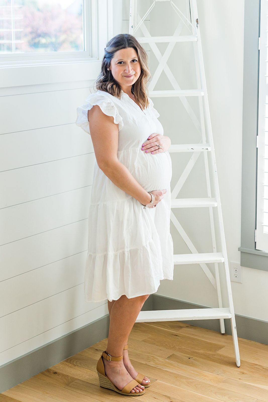 A mom to be in a white dress stands by a window in a studio holding her bump lactation consultant columbus ohio
