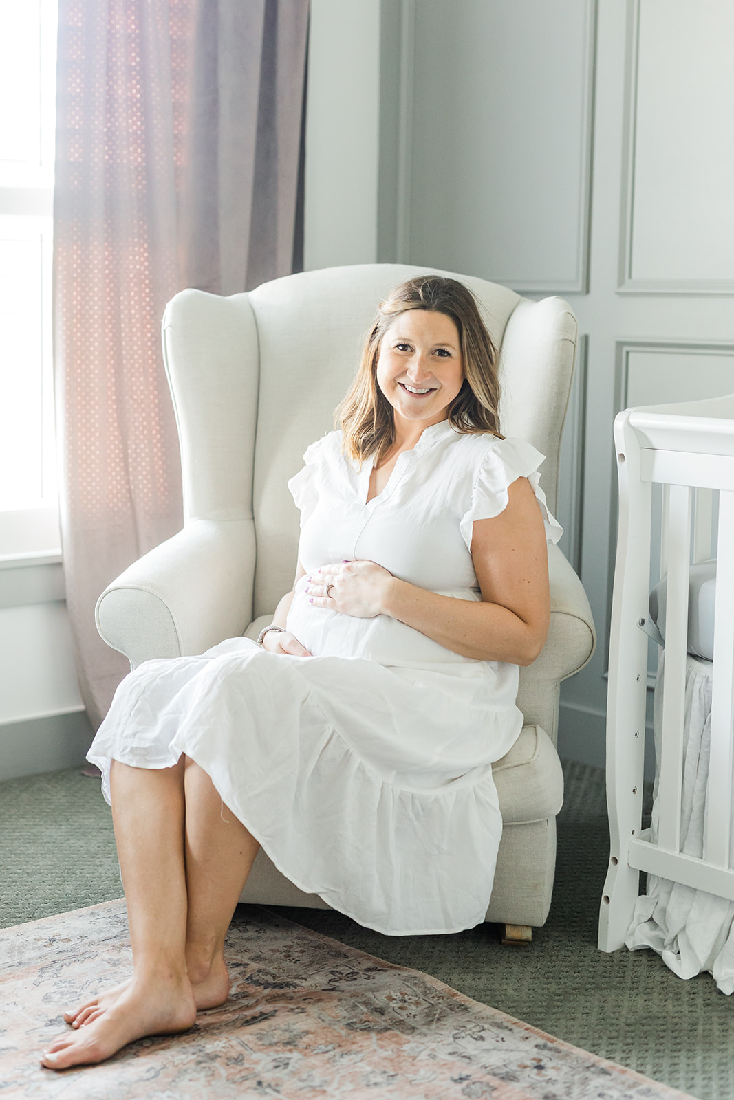 A mother to be in a white maternity dress sits in a nursing chair with hands on her bump lactation consultant columbus ohio